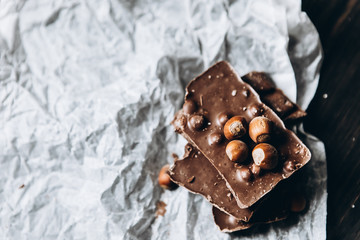 Milk chocolate with nuts: peanuts, hazelnuts on white parchment paper with a nutcracker and on a dark wooden table. Selective focus