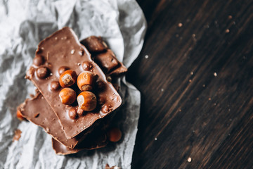 Milk chocolate with nuts: peanuts, hazelnuts on white parchment paper with a nutcracker and on a dark wooden table. Selective focus