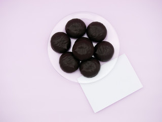 Chocolate-covered marshmallows on a transparent plate, An empty list      for recording (note)  on a pink background. Mockup.  The view from the top. 