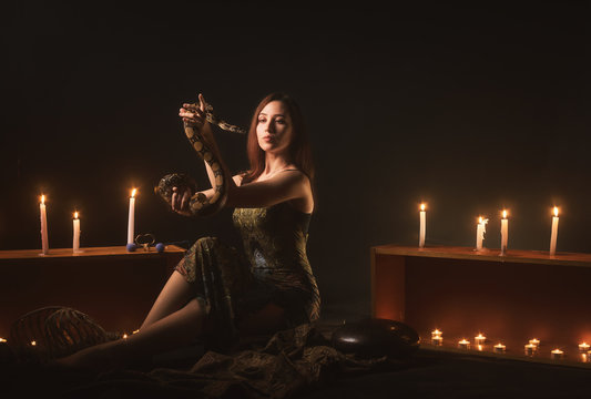 a witch with a snake on her neck and in her hands sits in a haze against a background of candles and a skeleton