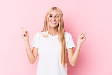 Young blonde woman on pink background indicates with both fore fingers up showing a blank space.