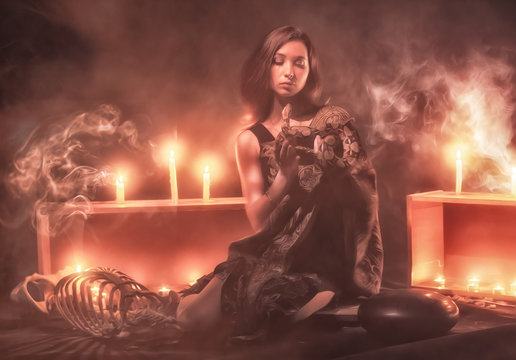 a witch with a snake on her neck and in her hands sits in a haze against a background of candles and a skeleton