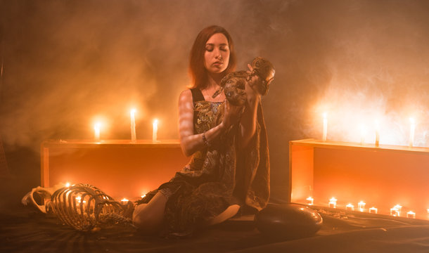 a witch with a snake on her neck and in her hands sits in a haze against a background of candles and a skeleton