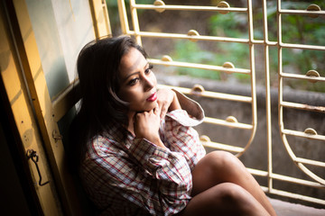 An young and attractive Indian  brunette woman in long western checkered shirt sitting on a window/balcony. Indian lifestyle.