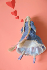 Funny little pocket doll made of fabric in flying skirts and with long linen braids on a coral background. The doll confesses in love.