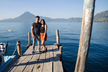 Fototapeta na wymiar Traveling couple standing at the dock of Lake Atitlan smiling at the camera - sunrise on Lake Guatemala - Hispanic adventure couple in Latin America tourist destination