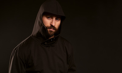 A man in a black tracksuit on a black background looks confidently into the frame. A late portrait of a guy with a haircut and beard dressed in a hood. Calm and strong look.