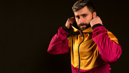 A man on a black background puts on the hood of a tracksuit. The modern image of an athlete with a beard and a beautiful hairstyle. The confident look of a strong man