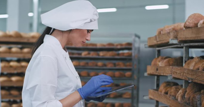Pretty young lady baker make some pictures with her tablet of a fresh baked bread from the shelves then she check the photos ,wearing white uniform with hat in a bakery industry