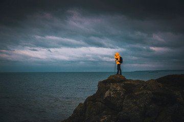 A young man adventurer with contrast yellow jacket posing over cliff by sea under gloomy sky with dramatic clouds