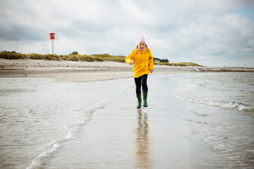 Cheerful little girl running on water of Baltic sea in rubber boots at windy weather