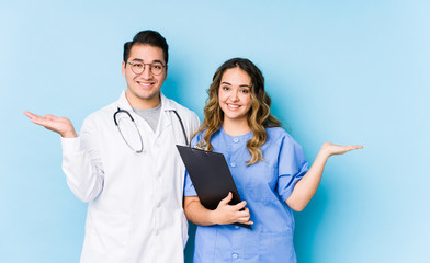 Young doctor couple posing in a blue background isolated makes scale with arms, feels happy and confident.