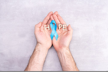 Health care and medicine concept - blue prostate cancer awareness ribbon in the hands of a man and the word hope, achalasia and adrenocortical cancer, on a gray background, flat lay