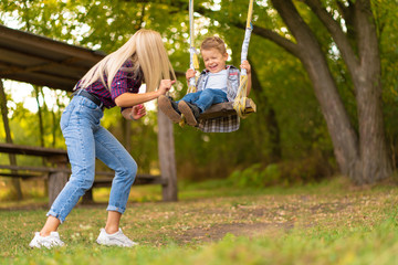 Young blonde mom shakes her little son on a swing in a green park. Happy childhood.