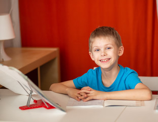 Fototapeta na wymiar smiling pretty fair-haired blue-eyed boy 9, a schoolboy, years old does homework, homework at the table, red background
