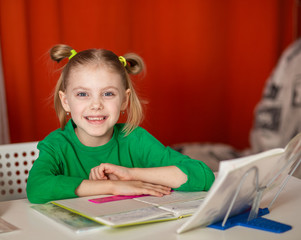 little cute smiling girl of 7 years old, first grader, left-hander does homework, writes in a notebook, red background, green jacket