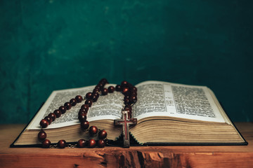 Open Holy Bible and beads crucifix on a red old wooden table. Beautiful green wall background.