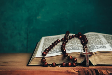 Close up Open Holy Bible and beads crucifix on a red old wooden table. Beautiful green wall background.