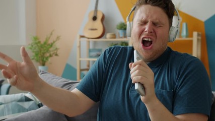Emotional young man is singing in remote control wearing wireless headphones enjoying song indoors at home. Modern devices and entertainment concept. - Powered by Adobe