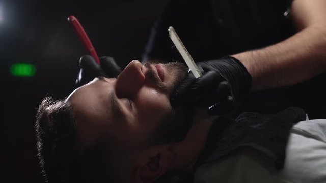 Male barber in black gloves shaving bearded client