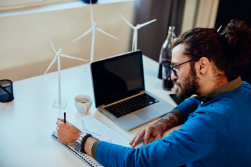 Dedicated innovative caucasian engineer sitting in his office and drawing windmill. Sustainable development concept.