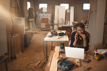 Young woman worker in the carpenter workroom. Woman making notes standing at table in modern...