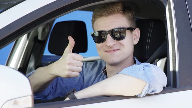 Young man gives thumb up in his car. Handsome brunette guy cool face in a car window. Blue screen chroma key background. Life winner fancy successful businessman making a face and thumbs up. Brunette
