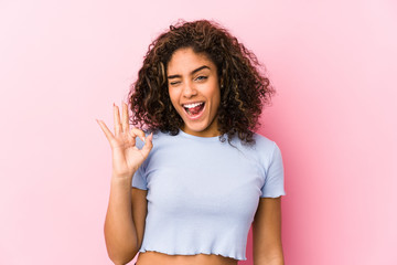 Young african american woman against a pink background winks an eye and holds an okay gesture with...