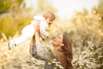 A beautiful young, happy woman with a baby in the spring in a blooming garden, smiling and tossing the baby. Concept: spring. Mother's day. Relationships in the family. March 8.