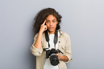 Young african american photographer woman holding a camera pointing his temple with finger, thinking, focused on a task.