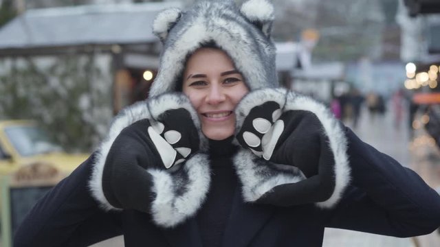Portrait of cheerful Caucasian woman in wolf hat and gloves smiling and waving at camera. Happy young girl spending weekends on winter fair. Leisure, resting, lifestyle.