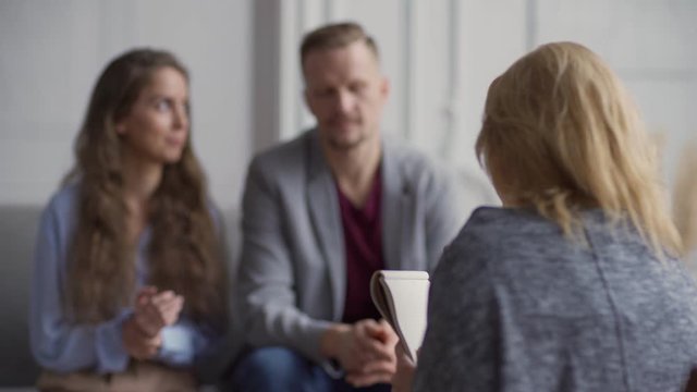 Professional female psychologist with notebook in hands having meeting with adult married couple and explaining details of interview and psychotherapy session