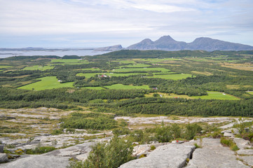 Fototapeta na wymiar View to green valley of Alsten island from hiking trail going through grey granite cliffs of Seven Sister mountains against the background of Donna island and blue Norwegian sea on sunny summer day