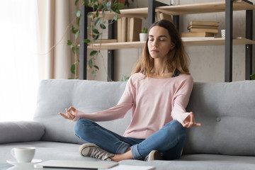 Millennial woman meditating in lotus pose in living room.