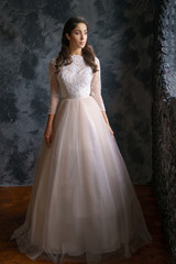 Beautiful young woman stands in a delicate wedding dress against a dark wall