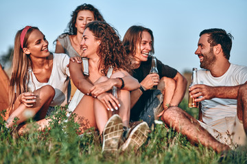 Five happy friends spending free time together in park sitting on grass, drinking beer and...