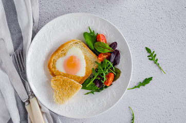 Piece of bread toast cut in shape of heart with egg on a white plate with arugula and cherry tomatoes. Romantic breakfast for Valentine's Day. Top view. Horizontal orientation.