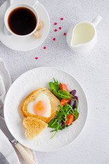 Piece of bread toast cut in shape of heart with egg on a white plate with arugula and cherry tomatoes. Romantic breakfast for Valentine's Day. Top view. Vertical orientation.