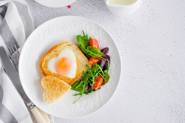Piece of bread toast cut in shape of heart with egg on a white plate with arugula and cherry tomatoes. Romantic breakfast for Valentine's Day. Top view. Copy space. Horizontal orientation.