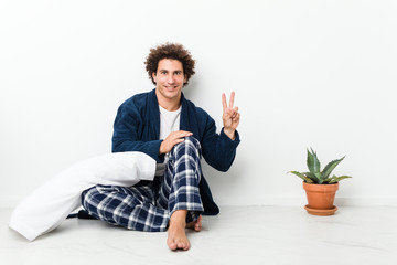 Mature man wearing pajama sitting on house floor showing victory sign and smiling broadly.