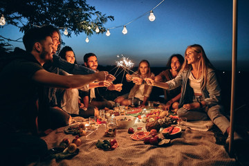 Happy friends having fun with fire sparkles. Young people millennials camping at picnic after...