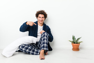 Mature man wearing pajama sitting on house floor person pointing by hand to a shirt copy space, proud and confident