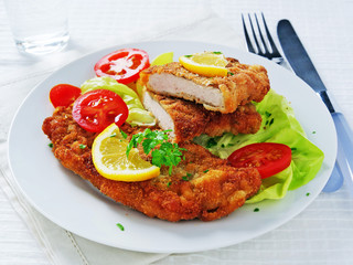 Wiener schnitzel over lettuce leaves with sliced tomatoes and lemon on white plate