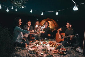 Group of young happy friends having picnic outdoors with sparklers.
