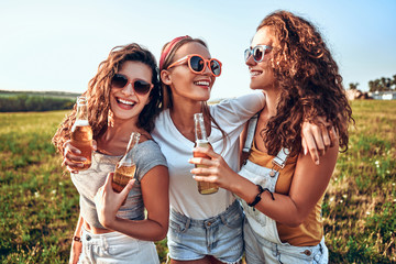 Candid of young attractive three girl group of friends cheers holding a bottle beer drinking for celebration party in evening sunset