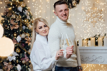 Obraz na płótnie Canvas Loving couple near the Christmas tree at home. Man and woman hug each other tender standing before a Christmas tree. Beautiful young couple is holding glasses of champagne
