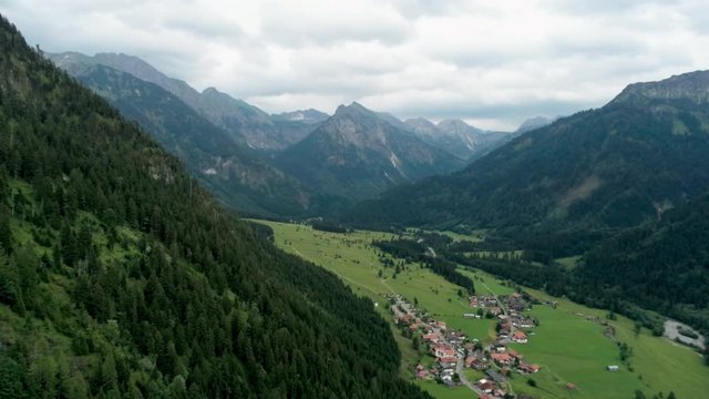 Majestic Aerial View of Village in Alps Mountains. Alpine Countryside with River, Forest and Car road. Agriculture and Ecotourism in Hinterstein, Germany, Europe. 4K Drone Pull in Shot