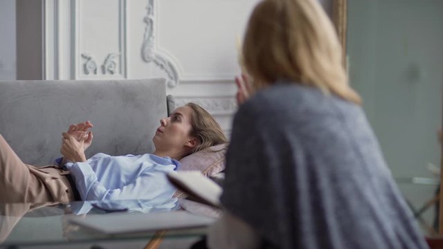Tracking left shot of nervous young woman lying on sofa and talking about her problems with defocused female therapist