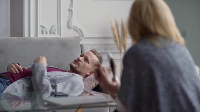 Tracking left shot of puzzled middle aged man in casual wear with clasped hands lying on sofa talking about problems with female psychologist at appointment