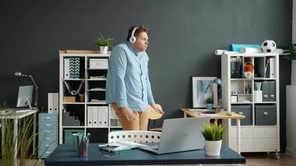 Slow motion of funny employee attractive guy dancing in office room wearing headphones having fun alone. Modern gadgets, joy and entertainment concept. - Powered by Adobe